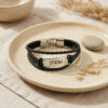 Black braided leather bracelet with a silver nameplate engraved with a Hebrew name, displayed on a ceramic plate