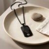 Black dog tag necklace with Hebrew engraving coiled in a ceramic dish, a men's stainless steel Judaica pendant