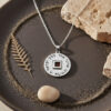 Silver Shema Yisrael nano Bible necklace on a dark stone tray with sand, a pebble and a gold leaf