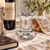 Crystal kiddush cup on a Shabbat table with a wine bottle, challah cover, candlestick and prayer book