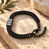 Black Hebrew name bracelet for dad with three engraved name beads on a wheat chain, resting on travertine stone with an olive branch