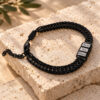 Black engraved family bracelet for dad on travertine stone beside an olive branch, three name beads