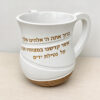 Angled view of the white Netilat Yadayim washing cup with the handle on the right, gold Al Netilat Yadayim blessing and brown base
